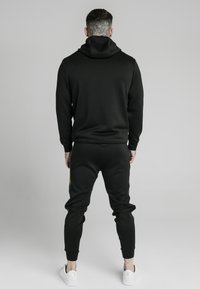 Czarna bluza z kapturem i dopasowane dresy wykonane z gładkiego materiału, z wysokim kołnierzem i dopasowanymi mankietami. Sneakery są białe z minimalistycznym wykończeniem.