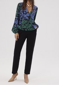 Blouse à manches longues en noir, vert et bleu à motif floral, associée à un pantalon noir et des escarpins beiges à bout pointu. Présente un décolleté en V profond.