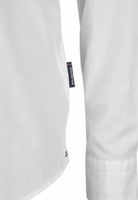 Witte shirt met een gladde, gestructureerde stof, met een rond afgewerkte onderkant en een zichtbaar merklabel aan de zijkant. De manchetten zijn keurig afgewerkt.