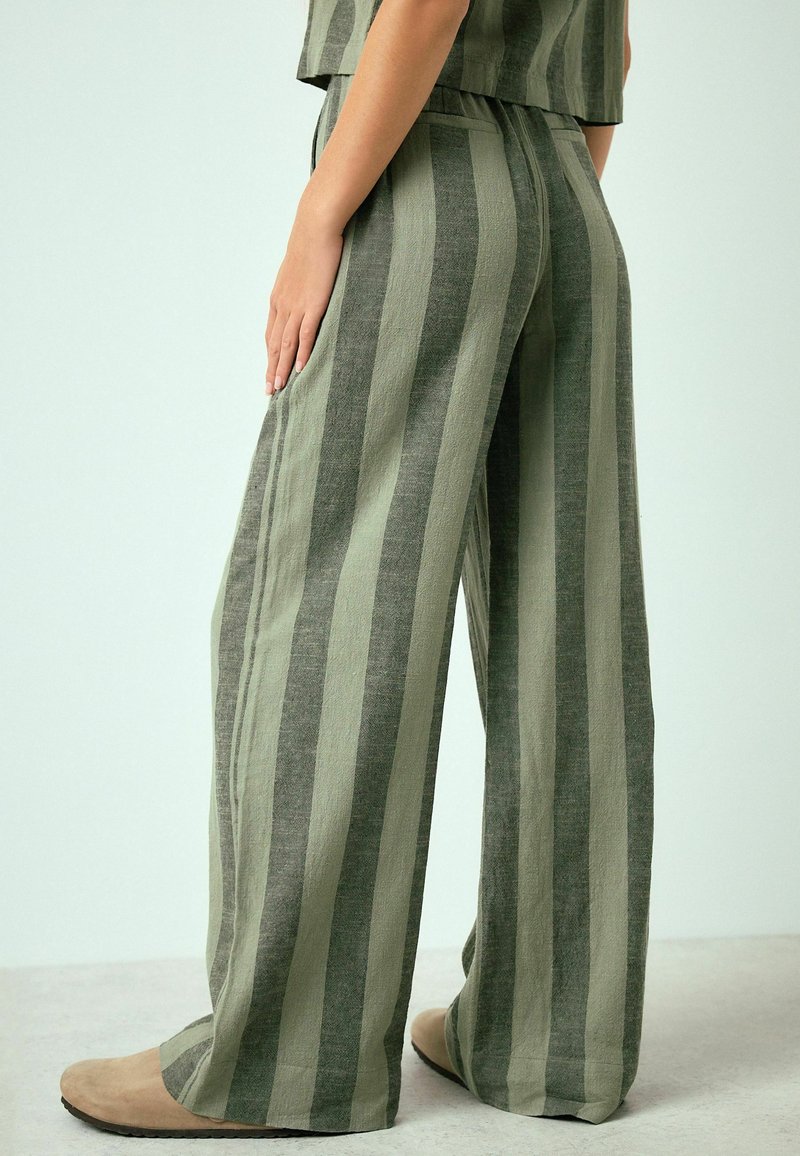 Persona que lleva pantalones anchos de pierna ancha y holgados con rayas verticales verdes y grises, combinados con zapatos planos beige.