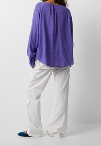 Top de manga larga morado en tela ligera, con cuello redondo y corte relajado. Combinado con pantalones cargo blancos de pierna ancha y zapatos planos azules.
