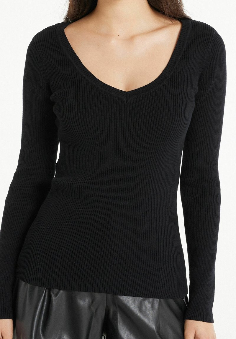Femme portant un haut noir côtelé ajusté à manches longues avec un décolleté en V et un pantalon en cuir noir, vue du cou aux hanches.