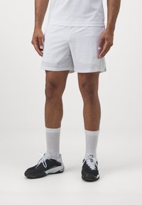 Pantalones cortos deportivos blancos hechos de un material ligero, con un logo sutil, combinados con zapatillas deportivas negras y calcetines blancos de caña corta.