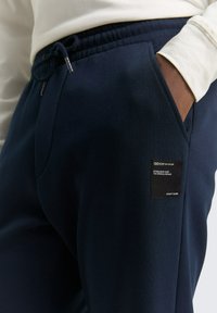 Granatowe spodnie dresowe wykonane z miękkiego materiału, z elastycznym pasem ze sznurkiem, bocznymi kieszeniami oraz czarną metką marki na lewej udzie.