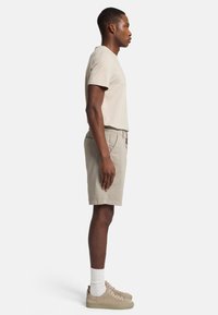 Beige katoenen t-shirt, lichtbeige shorts, gecombineerd met tan suède sneakers en witte sokken. Profielview benadrukt de casual pasvorm en het ontwerp.