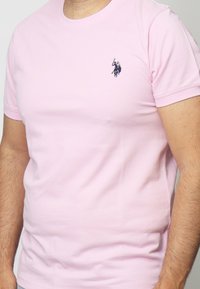T-shirt in cotone rosa chiaro con maniche corte, collo rotondo, dotata di un piccolo logo blu scuro nell'area sinistra del petto. Texture liscia.