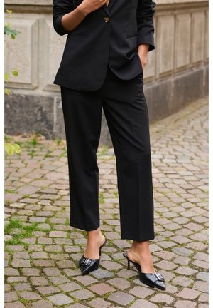 Person trägt schwarze, maßgeschneiderte Hose, passenden Blazer mit goldfarbenem Knopf und schwarze spitze High-Heel-Mules mit silberner Schnallendetail.