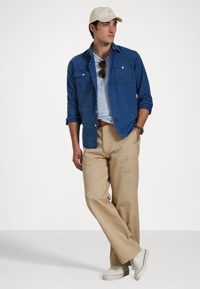 Lightweight denim shirt, light blue t-shirt, beige pants with distressed accents, white sneakers, and a cream cap with logo.