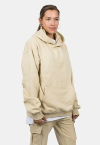 Beige hooded sweatshirt made from soft fabric, featuring a front pocket, loose fit, and ribbed cuffs. Worn over a white shirt.