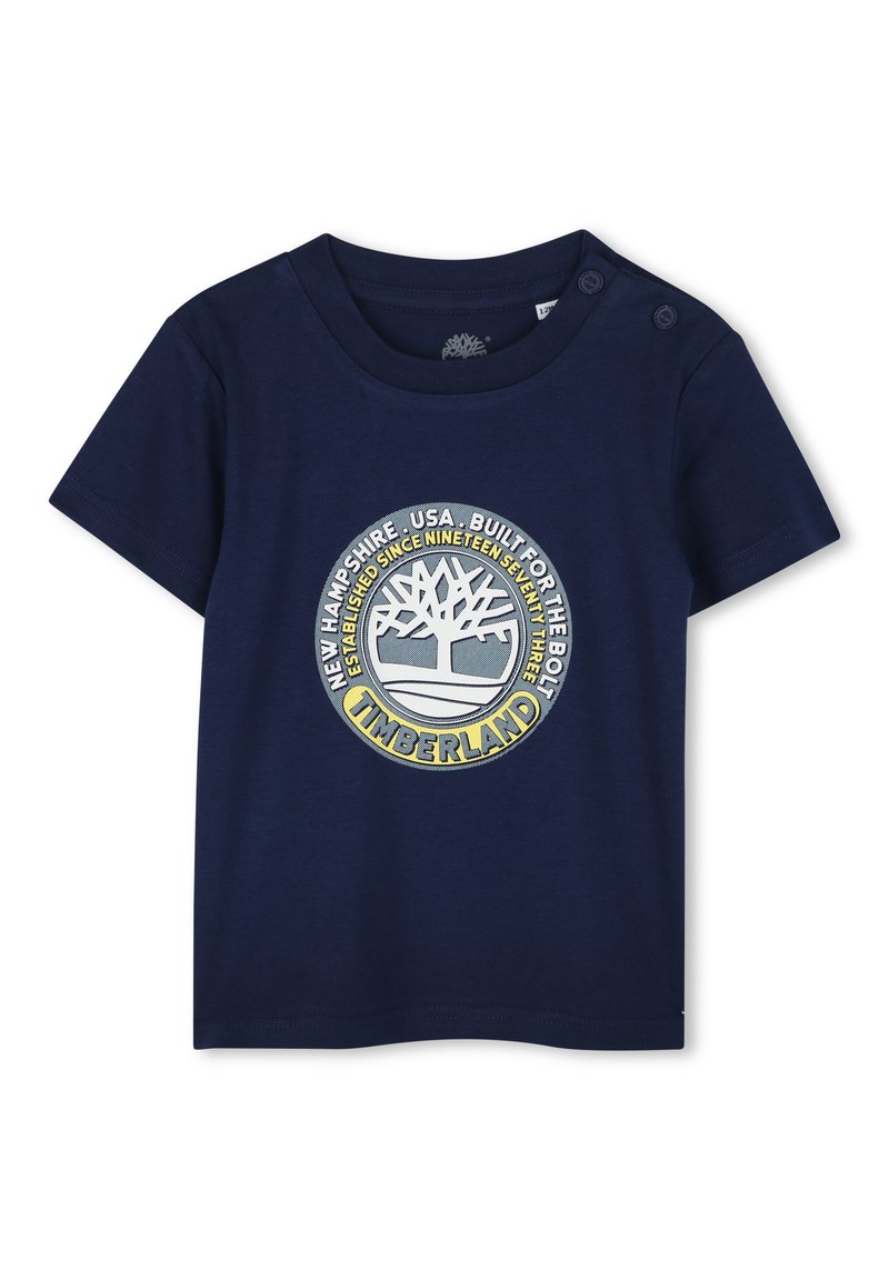 Camiseta de algodón azul marino con mangas cortas, que presenta un logo circular de Timberland en amarillo y gris, y botones a presión en el hombro.