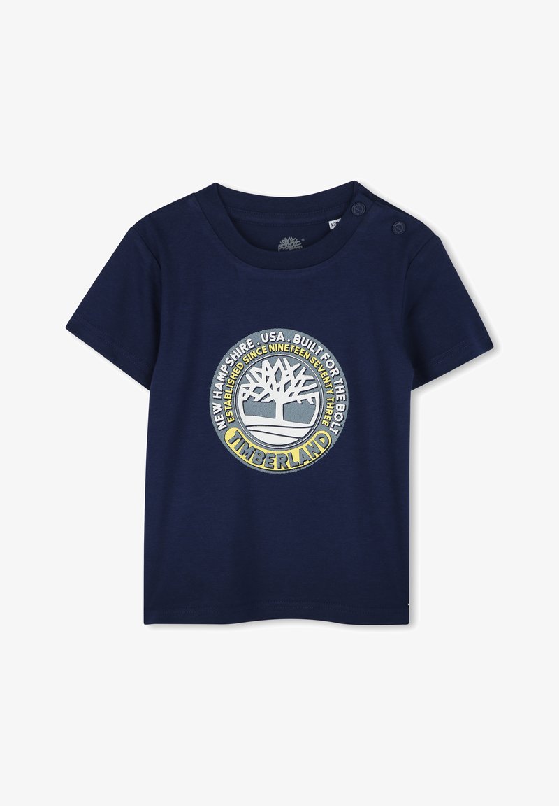 Camiseta de algodón azul marino con mangas cortas, que presenta un logo circular de Timberland en amarillo y gris, y botones a presión en el hombro.