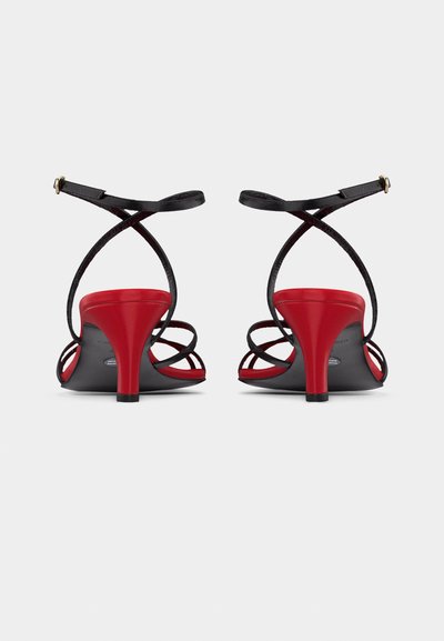 Ζευγάρι κόκκινων σανδαλιών με ψηλό τακούνι και λεπτά μαύρα λουράκια που σταυρώνονται στον αστράγαλο, εμφανιζόμενα από πίσω σε λευκό φόντο.
