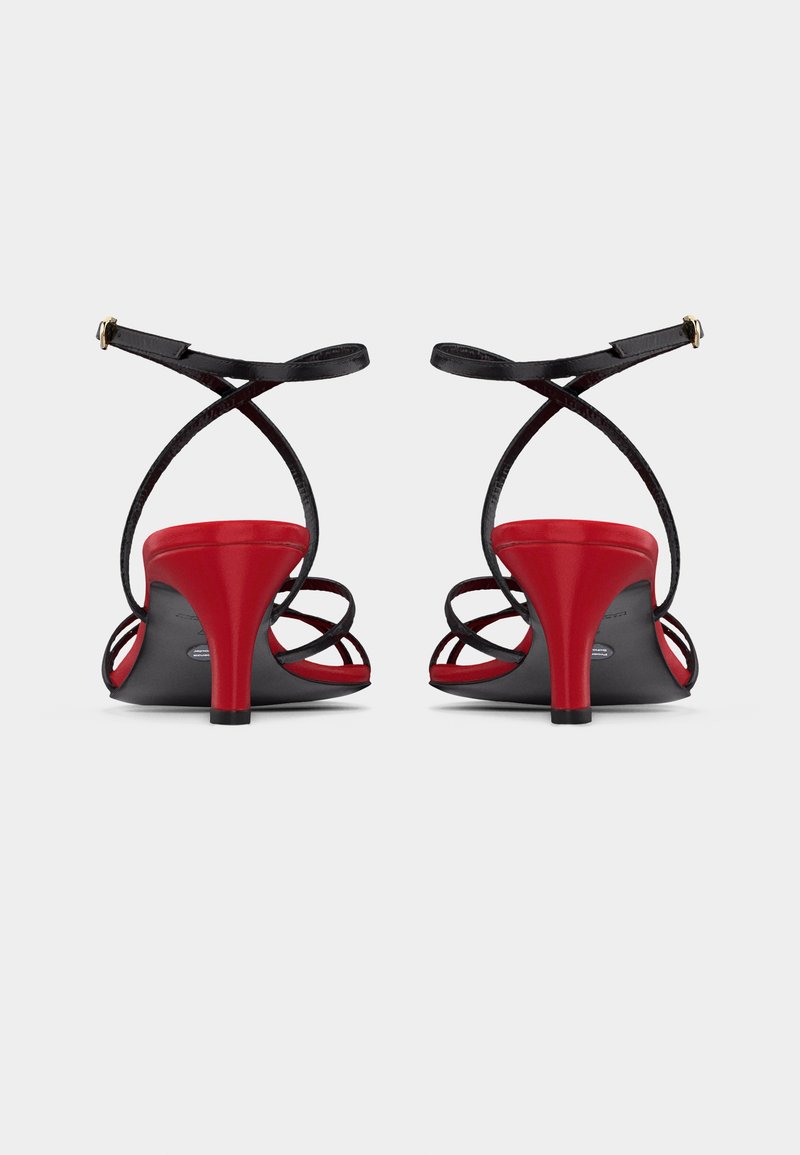 Paar rote Sandalen mit Absätzen und schwarzen, dünnen, sich kreuzenden Knöchelriemen, von hinten auf weißem Hintergrund gezeigt.
