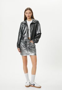 Giacca nera metallizzata con zip frontale, polsini elastici, abbinata a una gonna in denim argento lucido con spacco frontale e orlo sfilacciato. Top bianco e sneakers.