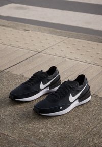 Fekete sportcipők sima bőr és hálós felsőrésszel. Fehér Nike emblémával, fekete fűzőkkel és kontrasztos fehér középtalppal.