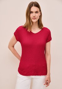 Top corto de manga corta texturizado en rojo, con escote redondo y dobladillo sutilmente curvado. Combinado con pantalones blancos de corte relajado.