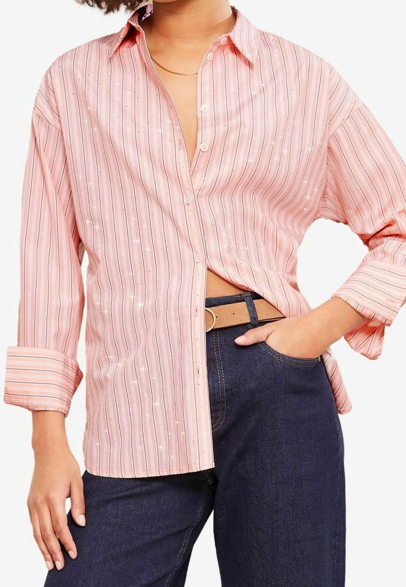 Personne portant une chemise ample à rayures roses avec boutons, manches retroussées, jean bleu foncé, ceinture beige et une main dans la poche.