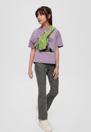 Meisje staat met een paarse Batgirl t-shirt, groene gebreide trui over de schouders, grijze jeans en witte platform sneakers, kijkend opzij.