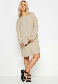 Abito a maniche lunghe beige con costine e scollo rotondo, che arriva sopra il ginocchio. Abbinato a sandali neri con cinturini e occhiali da sole tenuti in mano.