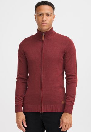 Maroon zip-up dukserica s rebrastim ovratnikom i manžetama; izrađena od pletenog materijala; ima malu oznaku brenda blizu rubnog šava.