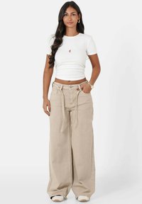 Witte, aansluitende t-shirt met een klein rood logo, gecombineerd met beige wijde broek met een trekkoord in de taille en zijzakken.