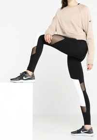 Leggings negros con acentos blancos y paneles de malla, combinados con un sudadera corta beige. Modelo lleva zapatillas de deporte con patrones en negro y gris.