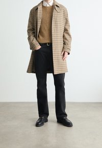 Beige- und braun karierter Mantel über einem braunen Strickpullover, kombiniert mit schwarzen Jeans und schwarzen Loafers, mit einem klassischen Kragen und großen Knöpfen.