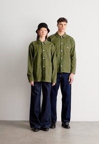 Δύο μοντέλα φορούν πουκάμισα σε ελιά πράσινη απόχρωση με κουμπιά και τσέπες στο στήθος, συνδυασμένα με σκούρα τζιν φαρδιάς γραμμής και μαύρα παπούτσια, σε ουδέτερο φόντο.