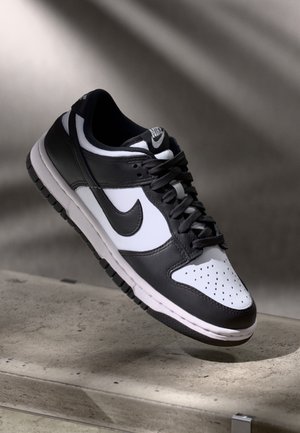 Schwarz-weißer Nike-Sneaker mit Schnürsenkeln, schwebend über einer Betonoberfläche vor einem verschwommenen grauen Hintergrund.