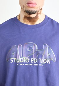 Violets krāsas kokvilnas T-krekls ar spīdīgu, lielizmēra "ALPHA" logo un "STUDIO EDITION" tekstu baltā krāsā. Tuvplāns par apkakles un dizaina detaļām.