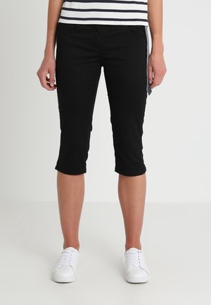 Pantalons capri noirs en tissu extensible, présentant une coupe slim, une taille mi-haute et une longueur au genou. Associés à des baskets blanches.