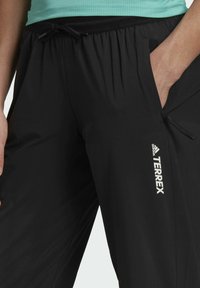 Pantalones deportivos negros confeccionados en material ligero. Cuentan con una cinturilla elástica con cordón, un bolsillo lateral y el logo "TERREX" en blanco.
