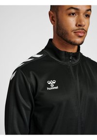 Svart sportjacka i slät tyg med hög krage, med vita chevronmönster på axlarna och en logotyp på bröstet.