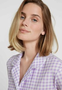 Paarse en witte gingham-gepatroniseerde pyjamajack met een ingezette kraag, zachte stof en subtiele glans. Het gezicht van het model is zacht belicht vanaf de zijkant.