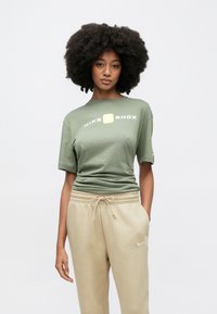 Πράσινο ελιάς κοντομάνικο μπλουζάκι Nike με το λογότυπο "NIKE SHOX" σε λευκό και κίτρινο χρώμα· φορεμένο με μπεζ φόρμα με κορδόνι στη μέση.