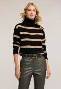 Černý a béžový pruhovaný rolák z měkkého materiálu, s volným střihem a žebrovanými manžetami, kombinovaný s olivově zelenými kalhotami a opaskem s cvočky.