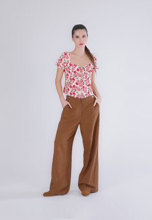 Femme debout les mains dans les poches, portant un chemisier blanc à motifs floraux avec des manches bouffantes et un pantalon large marron, sur un fond uni.