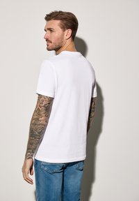 Bílý bavlněný tričko s krátkými rukávy, kulatým výstřihem a volným střihem. Nosené s vybledlými modrými džíny, které mají složité tetování na rukou.