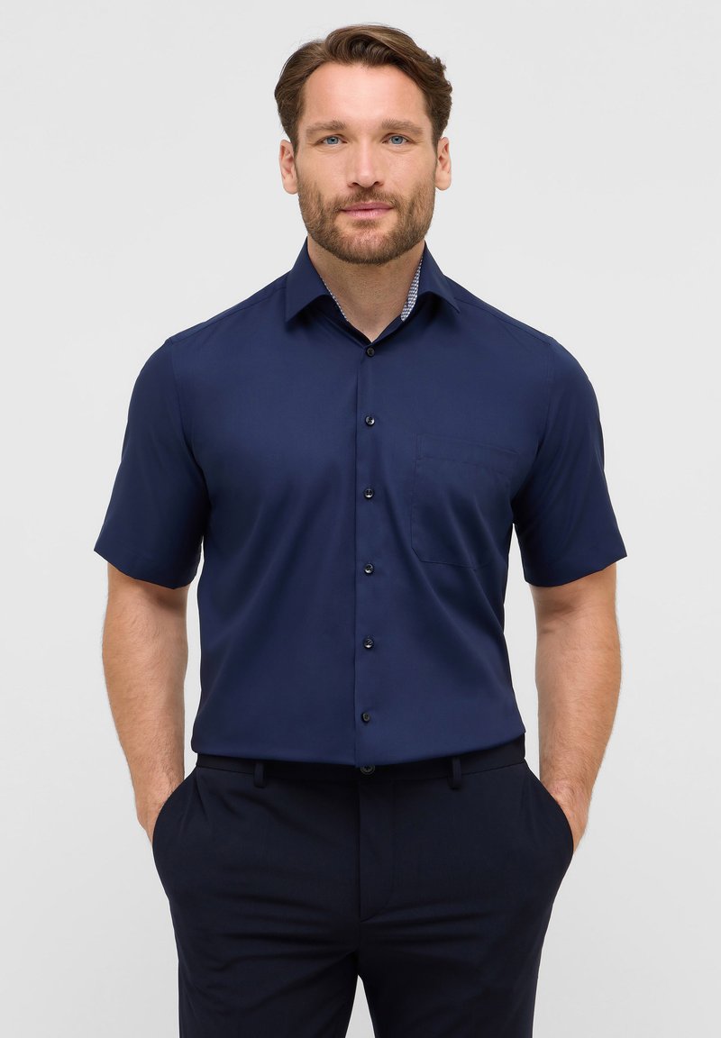 Camicia a maniche corte di colore blu navy, realizzata in tessuto liscio, con una tasca sul petto e dettagli a righe sotto il colletto.