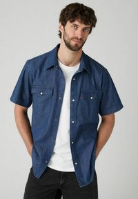 Camisa de mezclilla de manga corta en azul oscuro, con dos bolsillos en el pecho, un cierre de botones en la parte delantera y un cuello clásico. Llevada sobre una camiseta blanca.