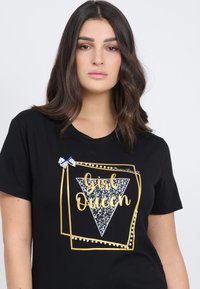 Camiseta negra de manga corta con un gráfico dorado y blanco que dice "Chica Reina" en cursiva, acentuado por un lazo de lunares azules y detalles de perlas.