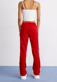 En person står med ryggen vänd mot betraktaren, klädd i en vit cropped tanktop, röda träningsoverallbyxor med sidostlister och vita sneakers på ett blått golv.