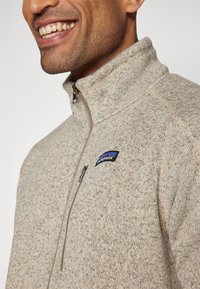 Giacca in pile beige con colletto alto, dotata di chiusura con zip e un piccolo logo Patagonia sul lato sinistro del petto. Tessuto strutturato.