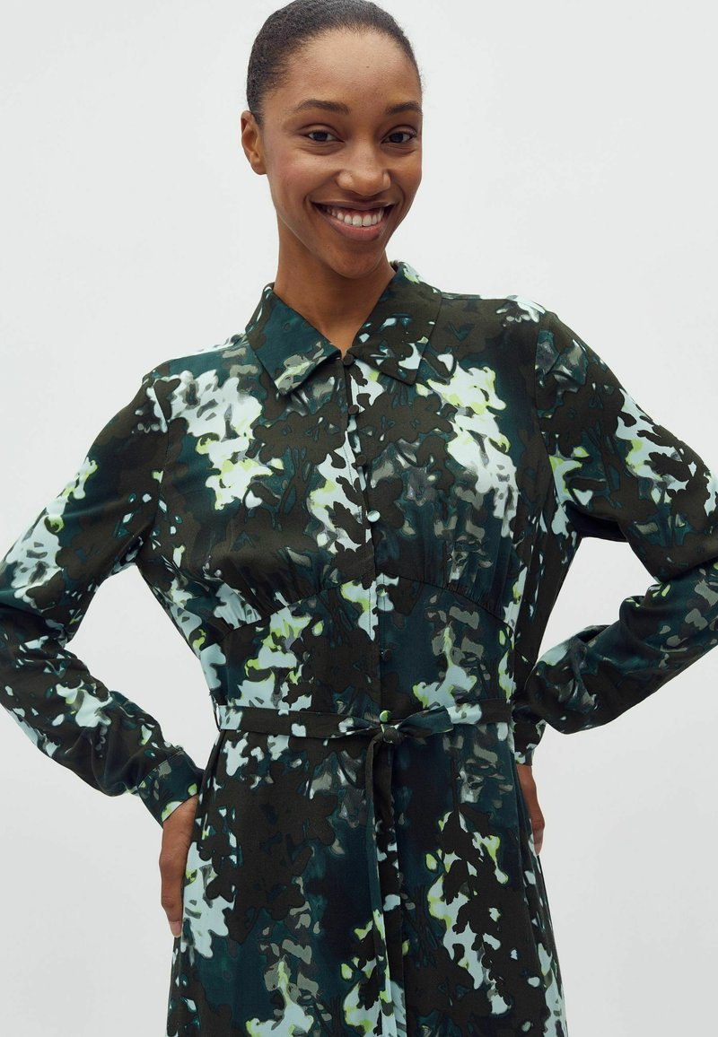 Personne souriante portant une robe à manches longues avec un col, à motif floral abstrait noir, vert, blanc et jaune, debout les mains sur les hanches.