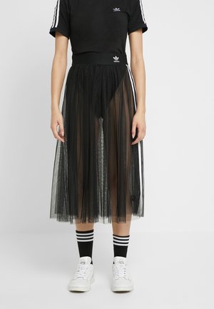 Persoon draagt een zwart Adidas T-shirt en een doorzichtige zwarte tule rok met Adidas-logo, zwarte gestreepte sokken en witte sneakers, staand tegen een witte achtergrond.