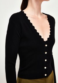 Donna in un cardigan nero aderente con bordi scallop e bottoni dorati, abbinato a pantaloni verde oliva su uno sfondo semplice.