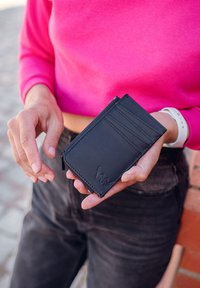 Black leather wallet with card slots and a zipper pocket, featuring clean lines and a textured finish, held in hands against a pink top.