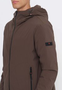 Mann trägt eine braune Kapuzenjacke mit schwarzem Reißverschluss und Reißverschlusstasche, steht vor einem schlichten weißen Hintergrund.