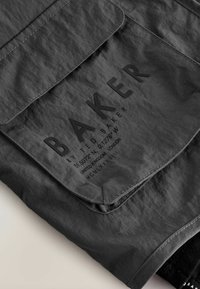 Szara torba typu worek wykonana z tkaniny, z dużym, wytłoczonym logo "BAKER", przeszyciami oraz zapięciem na klapkę. Zawiera szczegóły tekstowe.