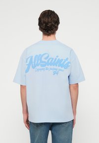 Άτομο με κοντά σγουρά μαλλιά που φοράει ένα ανοιχτό μπλε t-shirt AllSaints με το κείμενο «Απολαμβάνοντας το ταξίδι από το '94» στην πλάτη και μπλε τζιν.